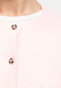 Κοντινό πλάνο ενός ατόμου που φοράει ένα ανοιχτό ροζ πλεκτό ζακέτα με μαύρα και λευκά μάρμαρα κουμπιά πάνω από ένα λευκό ριμπ πλεκτό τοπ.