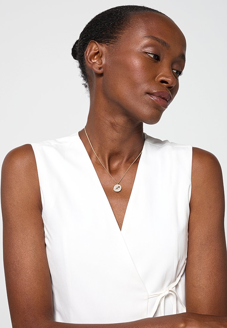 Biały top w formie owinięcia, wykonany z gładkiego materiału, z dekoltem w kształcie litery V i detalem w postaci wiązania w talii, w połączeniu z prostą złotą naszyjnikiem z zawieszką.