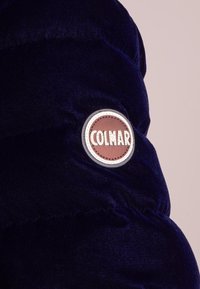 Mörkblå pufferjacka med en strukturerad yta och en rund brun tyglogotyp med ordet "COLMAR."