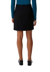 Zwarte rok met een getailleerde snit, tot de knie, met een ritssluiting aan de achterkant, een gladde textuur en minimale stikseldetails. G gedragen met zwarte schoenen.