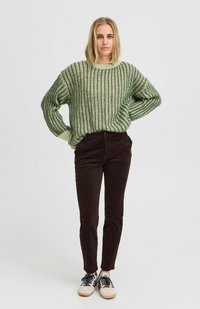 Grüner gestrickter Pullover mit geripptem Struktur, kombiniert mit dunkelbraunen Cordhosen. Die Schuhe sind helle Sneaker mit schwarzen Akzenten.