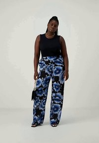 Črn brezrokavnik v kombinaciji z širokimi hlačami, ki imajo modri cvetlični vzorec na črni podlagi. Model drži majhno črno torbico.