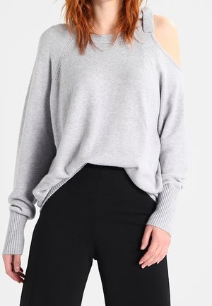 Maglione grigio chiaro con scollo off-the-shoulder, con polsini e orlo a costine. Presenta un dettaglio con fascia laterale, una texture morbida e una vestibilità rilassata. Abbinato a pantaloni neri.