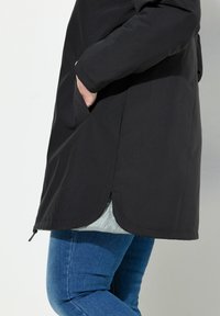 Schwarzer langer Mantel mit matter Oberfläche, ausgestattet mit einer verdeckten Reißverschlusstasche und abgerundetem Saum. Getragen über einem hellgrauen Innenfutter und blauen Jeans.