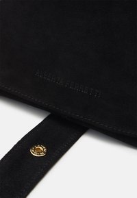 Sac en daim noir avec un bouton en métal doré, un logo embossé et un détail de couture contrastant sur la sangle. Texture lisse.