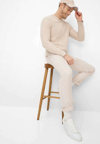 Mann trägt beigen Pullover, Hose und Kappe, sitzt auf einem Holzstuhl, richtet die Kappe und lächelt vor weißem Hintergrund.