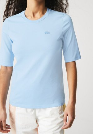 Frau trägt ein hellblaues Kurzarmhemd mit einem kleinen gestickten Logo auf der linken Brust und weiße Shorts mit Farbakzenten.