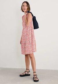 Rochie fără mâneci cu un model floral roșu și alb, cu un croi evazat, asortată cu o geantă bleumarin și sandale negre cu curele, pe un podea deschisă.