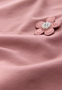 Rožnata tkanina z gladko teksturo, okrašena z vezenim apliciranim cvetom s perlno sredino in detajli na cvetnih listih.