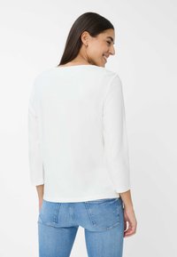 Weißes Langarmoberteil mit rundem Ausschnitt, glatter Textur und lockerer Passform, kombiniert mit hellblauen Jeans aus Denim mit Rückentaschen.