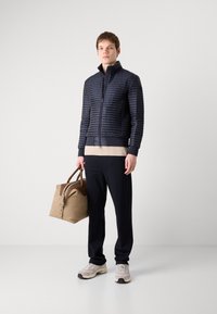 Doudoune marine avec texture matelassée, poignets et ceinture noirs, pull beige en dessous, pantalon noir et sac de sport beige.