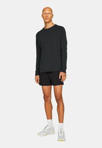 Svart långärmad skjorta i kombination med svarta shorts. Modellen bär vita och grå sportskor med detaljer, stående mot en enfärgad bakgrund.