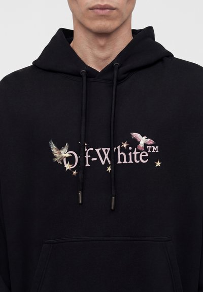 Άντρας που φοράει μαύρο hoodie με το κείμενο «Off-White», δύο πουλιά και σκορπισμένα αστέρια κεντημένα στο στήθος.
