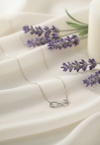 Silvrig oändlighetshalsband på ett delikat kedja, mot mjuk krämfärgad tyg, med lavendelflower i närheten som ger en touch av färg.