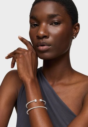 Primo piano di una persona che indossa una canotta grigia e due braccialetti d'argento, uno con ciondoli a forma di cuore e orsetto, posato davanti a uno sfondo semplice.