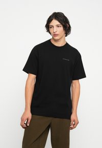 Zwarte katoenen t-shirt met ronde hals en korte mouwen, voorzien van een klein grijs logo op de voorkant. Draag het met olijfgroene corduroy broek.