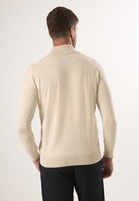 Beige pletený svetr s vysokým límcem a žebrovanými manžetami. Přiléhavý střih s dlouhými rukávy, který ukazuje hladkou strukturu a čisté provedení.