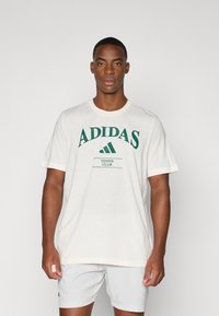 T-shirt de algodão branca com o logótipo verde "ADIDAS" e o texto "TENNIS CLUB". Mangas curtas com decote redondo e um corte descontraído.