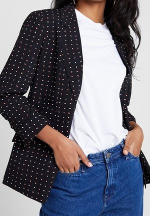 Femme portant un blazer noir à petits pois rouges et blancs sur un t-shirt blanc, associé à un jean en denim bleu, les mains dans les poches.