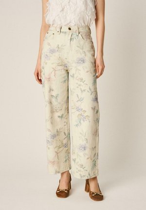 Mujer vestida con pantalones de tiro alto y pierna ancha en color crema con estampado floral pastel, combinados con una blusa blanca sin mangas con volantes y mocasines marrones con tacón.