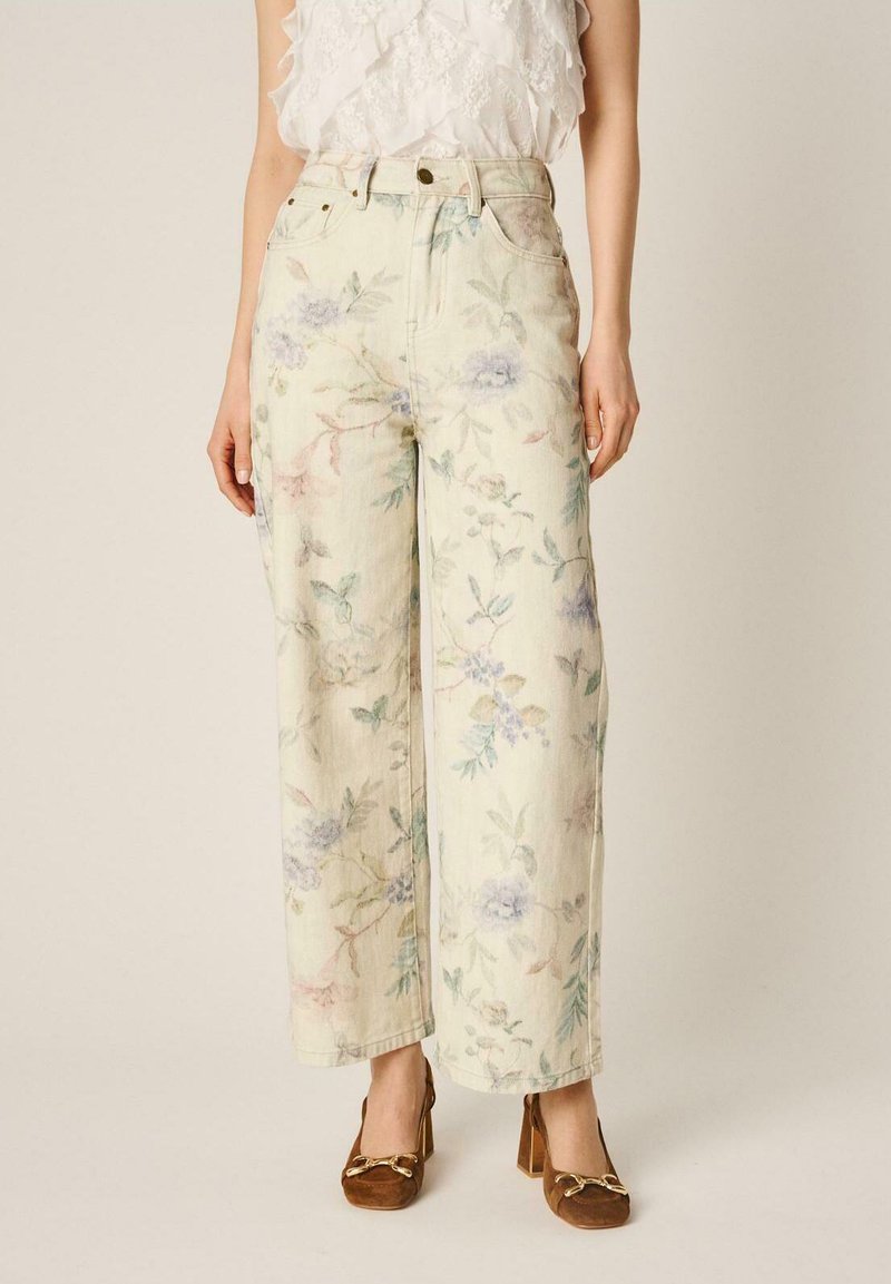 Mujer vestida con pantalones de tiro alto y pierna ancha en color crema con estampado floral pastel, combinados con una blusa blanca sin mangas con volantes y mocasines marrones con tacón.