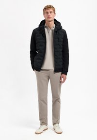 Zwarte pufferjack met een gestructureerde afwerking, capuchon en ritssluiting, gedragen over een lichtbeige poloshirt en beige broek, met witte sneakers.