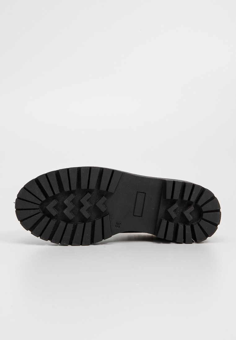 Semelle de botte en caoutchouc noir avec des motifs de crans profonds, présentant des motifs en zigzag pour la traction et une plaque rectangulaire au centre.