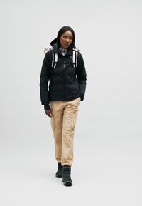Doudoune noire, pantalon cargo beige, bottes de randonnée noires et un sac à dos léger. Texture lisse, design pratique, avec des fonctionnalités ajustables.