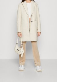 Abrigo de mezcla de lana color crema con dos botones negros, bolsillos laterales y un acabado texturizado. Pantalones de terciopelo beige y un pequeño bolso blanco roto con tachuelas.