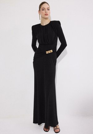 Kobieta stojąca na białym tle, ubrana w długą czarną sukienkę z poduszkami na ramionach i złotą klamrą w talii, oraz złote kolczyki.