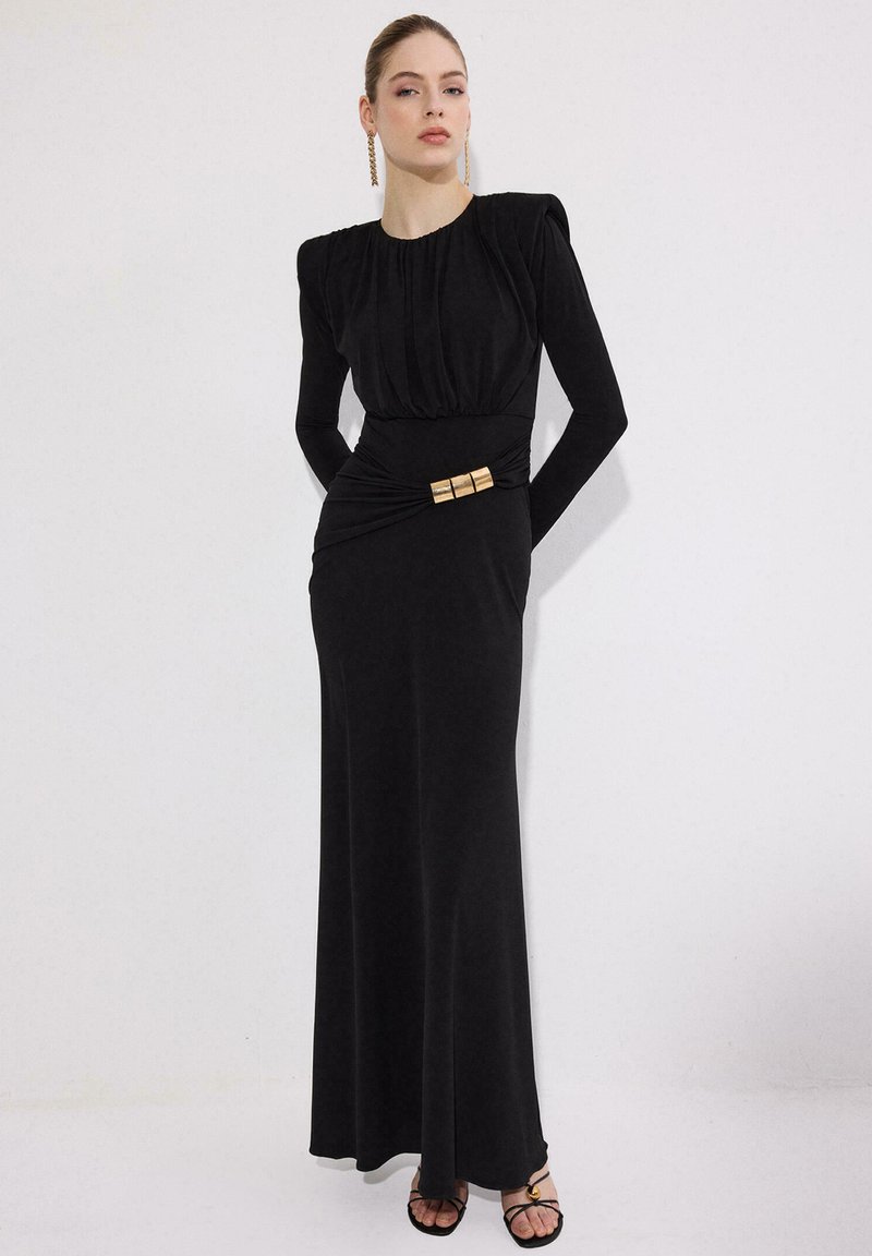 Γυναίκα που στέκεται μπροστά σε λευκό φόντο, φοράει μαύρο μακρύ φόρεμα με επωμίδες και χρυσή αγκράφα στη μέση, καθώς και χρυσά σκουλαρίκια.