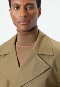 Olivengroene jas met scherpe revers over een bruine coltrui. Gladde stoftextuur, getailleerde pasvorm en subtiele stikseldetails op de jas.