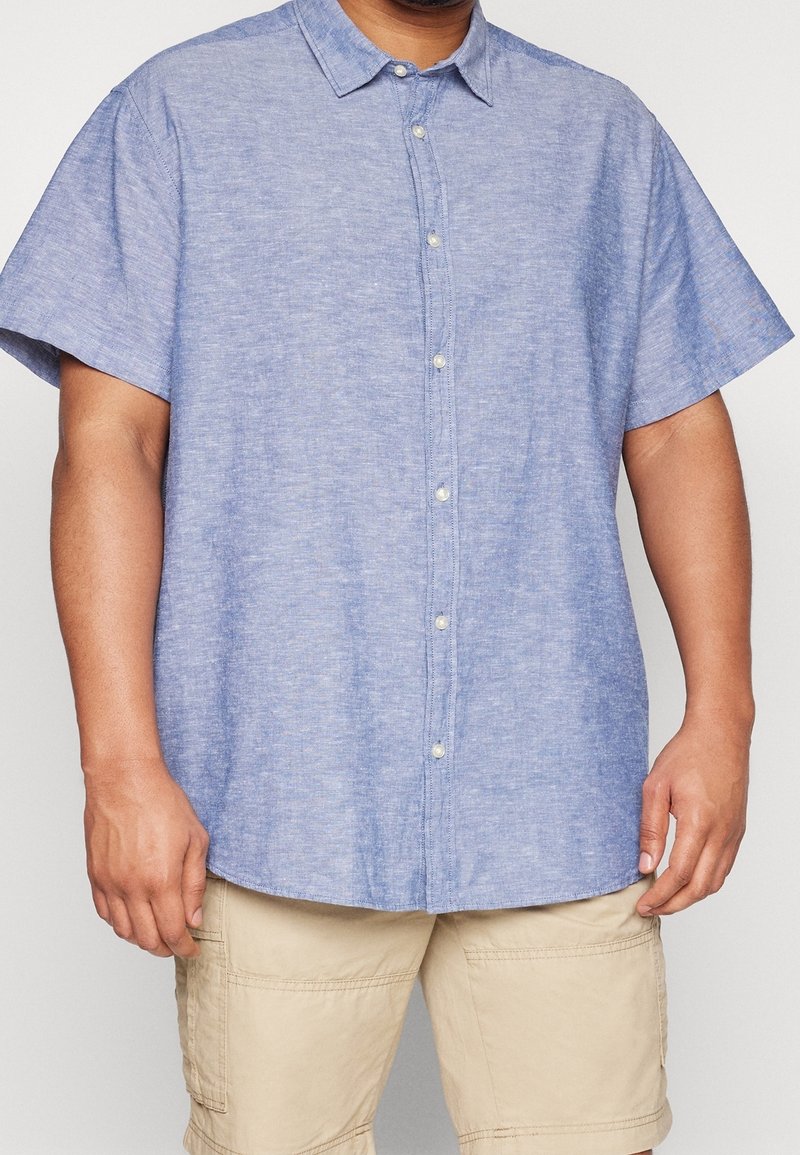 Krátký rukáv, světle modrá košile na knoflíky vyrobená z texturované látky s límečkem a bílými knoflíky, spárovaná s khaki šortkami.