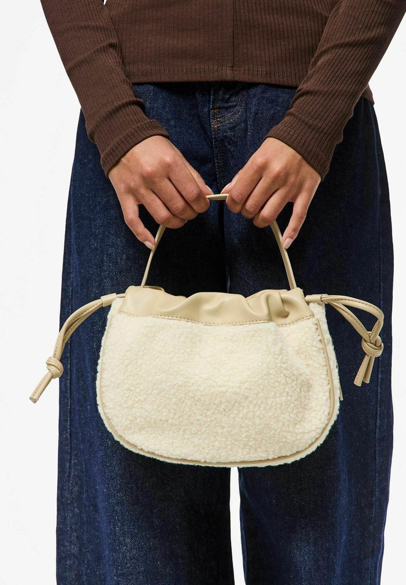 Cremefarvet håndtaske med en tekstureret, fluffy krop og en glat tan top. Har et samlet design og to knyttede håndtagsdetaljer.