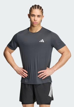 Giovane uomo con capelli intrecciati che indossa una t-shirt grigio scuro Adidas e pantaloncini neri con il logo Adidas, in piedi con le mani sui fianchi.
