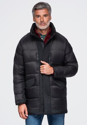 Homme d’âge moyen aux cheveux gris et à la barbe, portant une doudoune noire, une chemise à carreaux rouge et un jean bleu, debout devant un fond blanc.