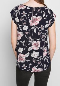 Femme portant un haut bleu marine avec des motifs floraux roses et blancs et une fermeture éclair visible dans le dos, associé à un pantalon noir.