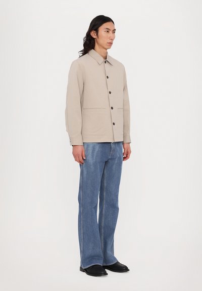 Jachetă bej cu guler, închidere cu nasturi și buzunare laterale, asortată cu blugi albaştri evazați și pantofi negri, pe un fundal neutru.