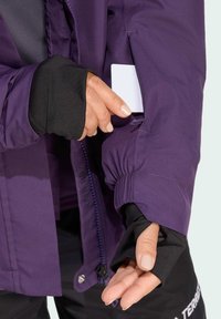 Violetti vedenpitävä takki, jossa on joustavat hihansuut, vetoketjullinen etuosa ja sivutasku. Mustat hihansuosat lisäeristystä varten.