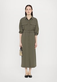 Olivgrünes, tailliertes Hemdkleid mit zwei Brusttaschen, Knopfverschluss und langen Ärmeln, kombiniert mit schwarzen Ballerinas und einer kleinen Tasche.