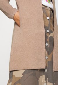 Cardigan en maille brun clair avec manches côtelées, comportant une large ouverture et des poches latérales, associé à une jupe à motifs avec boutons.