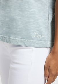Top azzurro chiaro a maniche corte con una texture morbida. Presenta un logo "Lee" ricamato in bianco sull'orlo. Indossato con pantaloni bianchi.