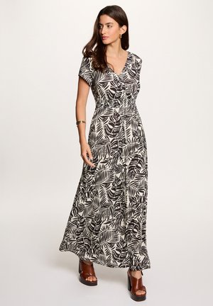 Vrouw die een zwart-witte maxi-jurk met bladpatroon en V-hals draagt, met een geplooide taille en bruine plateauhakken, staand tegen een effen achtergrond.