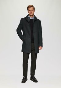 Donkergroene wollen jas met een knoopsluiting aan de voorkant, zijzakken en een capuchon. Gepaard met donkere broeken en zwarte leren laarzen.
