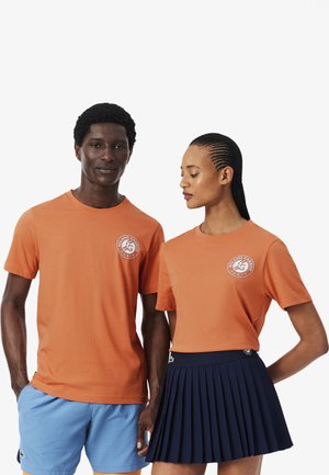 Homme et femme portant des t-shirts orange assortis Roland Garros ; homme en short bleu, femme en jupe plissée bleu marine, debout côte à côte.