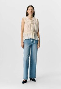 Blouse légère sans manches, plissée, avec un détail à nouer au niveau du col, associée à un jean bleu taille haute à jambe droite et des talons noirs.