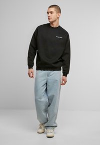 Sudadera negra con cuello redondo y un logotipo en la parte frontal, combinada con unos pantalones vaqueros holgados de color azul claro y zapatillas blancas. Textura de tejido suave.