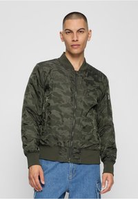 Veste bombardier camouflage vert foncé avec fermeture éclair devant, poignets et col côtelés. Comprend des poches latérales et un léger éclat sur le tissu.
