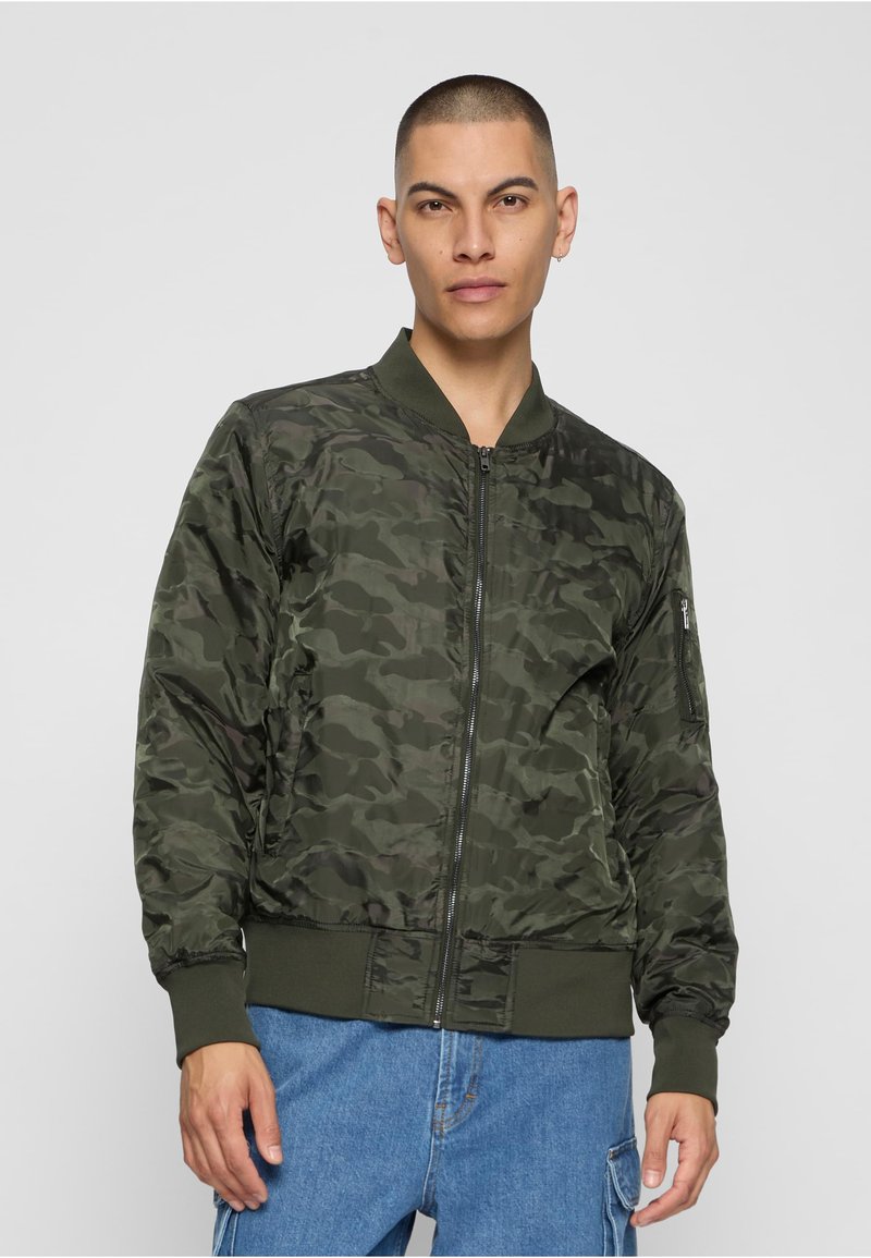Veste bombardier camouflage vert foncé avec fermeture éclair devant, poignets et col côtelés. Comprend des poches latérales et un léger éclat sur le tissu.