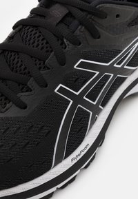 Chaussure de sport noire avec une tige en mesh, des lacets noirs et des accents de logo blanc bien visibles. Semelle texturée étiquetée "FlyteFoam."